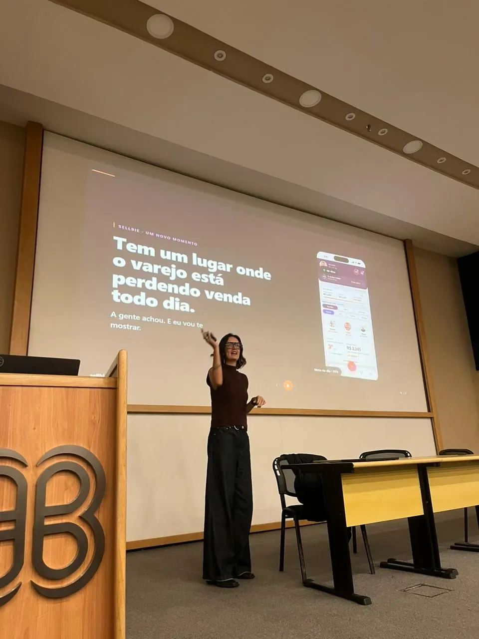 Jholy Mello apresentando em auditório, com os slides da palestra projetados ao fundo